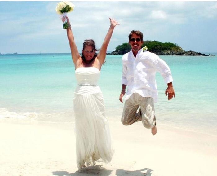 wedding couple, with bride holding wedding flowers and grooming jumping on beach, with the beautiful blue water of the United States Virgin Islands behind them